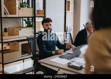 Bärtiger junger Mann in einem Rollkragen, der sich intensiv auf seine Laptop-Arbeit konzentriert, in einem modernen hölzernen Büro mit einem Kollegen in der Nähe. Stockfoto