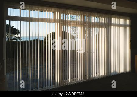 Sonnenlicht, das durch weiße vertikale Jalousien in voller Länge vor drei Glasschiebetüren strahlt, die zu einer Terrasse, Garten und Meer führen Stockfoto