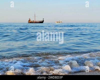 Zwei Seeschiffe trafen sich im blauen Meer – ein Schiff aus der Wikingerzeit und eine moderne Yacht. Die Wellen treffen auf die Steine, der ruhige, schöne wolkenlose Himmel. Beau Stockfoto