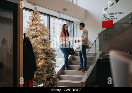 Zwei Kollegen, die ein freundliches Gespräch neben einem geschmückten Weihnachtsbaum in der Lobby eines modernen Bürogebäudes führen. Stockfoto