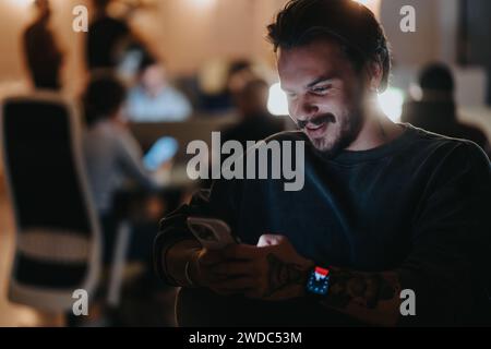 Ein zufriedener junger, erwachsener Mann greift mit seinem Smartphone auf und strahlt Glück aus in einem leise beleuchteten Abendbüro voller Kollegen. Stockfoto