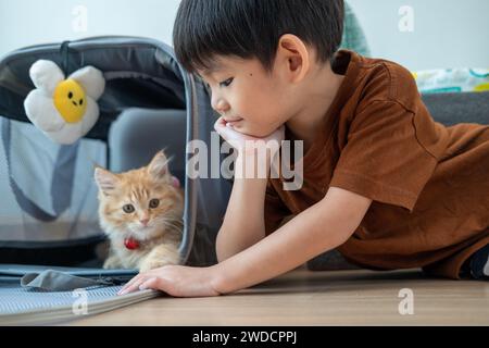 Ein kleiner asiatischer Junge sitzt und schaut liebevoll ein orangenes Kätzchen an. Stockfoto
