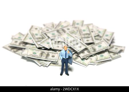 Miniatur-Kleinkinder-Spielfiguren-Fotografie. Anlageberatungskonzept. Ein Geschäftsmann, der vor dem Geldhaufen aus Papier steht. Isoliert auf einem weiß Stockfoto