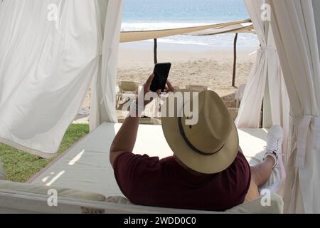 Dunkelhäutiger erwachsener Latino-Mann mit Sonnenbrille und Hut benutzt sein Handy, um Probleme auf einem Bett vor dem Strand im Urlaub zu lösen Stockfoto