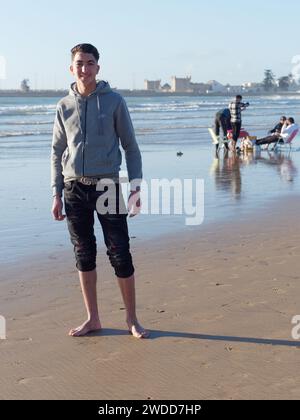 Ein hübscher junger Mann lächelt, als er am Strand steht, mit Leuten, die auf Liegestühlen sitzen, in Essaouira, Marokko, 19. Januar 2024 Stockfoto
