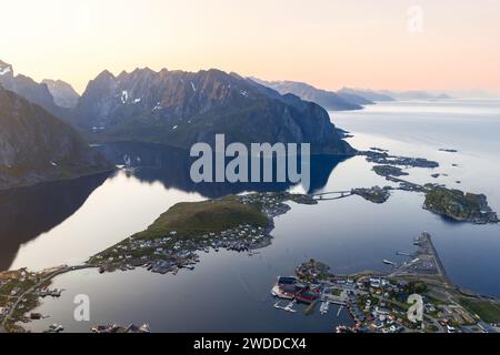 Aus der Vogelperspektive auf das ruhige reine-Dorf in Lofoten, Norwegen unter der Mitternachtssonne, mit Bergreflektionen auf ruhigen Gewässern, überbrückten Inseln Stockfoto