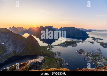 Atemberaubendes Panorama von Reinebringen mit der Mitternachtssonne, die hinter zerklüfteten Lofoten-Gipfeln eintaucht und ein warmes Leuchten über das reine Dorf wirft Stockfoto