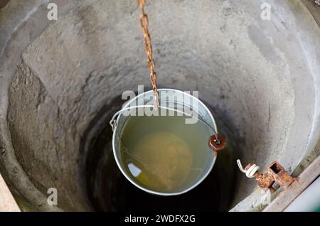 Metalleimer am Ziehbrunnen im europäischen Dorf. Retro Steinwasserbrunnen in ländlicher Umgebung Stockfoto