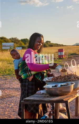 Dorfstraßenverkäufer, afrikanerin mit Baby im Rücken, verkauft Erdnüsse Stockfoto