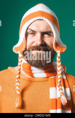 Nahaufnahme Porträt eines attraktiven Mannes mit Bart und Schnurrbart in orangefarbenem Pullover, lustigem Hut und gestreiftem Schal. Schöner bärtiger Mann in warmen Kleidern Stockfoto