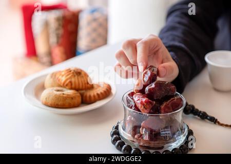 Hand mit eid-Süßigkeiten und Datteln auf weißem Tisch mit Rosenkranz und einer Tasse Kaffee Stockfoto