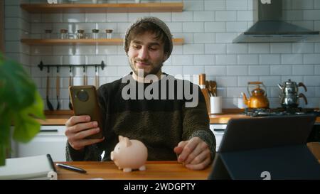 Glücklicher junger Mann mit Beanie, der zu Hause in der modernen Küche sitzt, während er das Smartphone in der Hand hält und auf die Kamera blickt und ein paar Münzen hineinsteckt Stockfoto