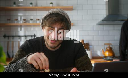 Junger Mann mit Beanie, der zu Hause in der modernen Küche sitzt, während er mit seinem Freund spricht, dreht sich nach vorne, steckt Münzen in die Schweinebank und schaut sich das an Stockfoto