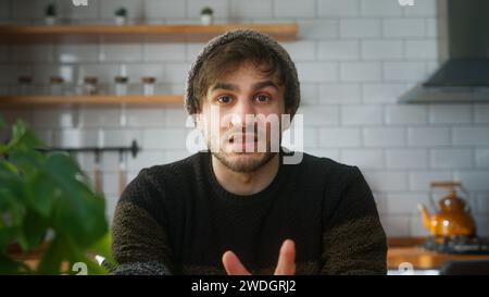 Junger erwachsener Mann mit Beanie, der in der modernen Küche zu Hause sitzt und auf die Kamera schaut, während er eine Konferenzschaltung hält, dem Lautsprecher beim Videoanruf zuhört. Stockfoto