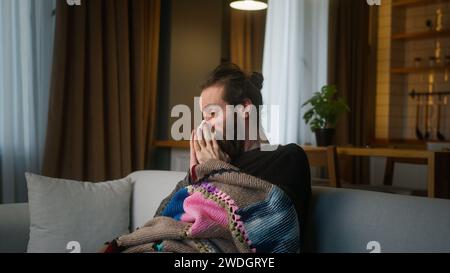 Kranker bärtiger Mann, der sich mit einer warmen Decke bedeckt, leidet an Husten und laufender Nase, während er auf dem Sofa in einem modernen Zuhause sitzt Stockfoto