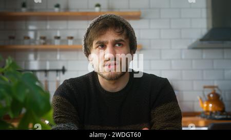 Junger Mann mit Beanie, der zu Hause in der modernen Küche sitzt und in die Kamera schaut, während er eine Konferenzschaltung hält und dem Lautsprecher beim Videoanruf zuhört Stockfoto