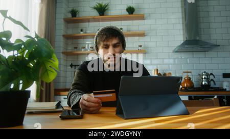 Junger Mann mit Beanie, der in der Küche zu Hause sitzt und die Kreditkartennummer auf dem Tablet-Gerät eingibt, macht eine sichere, einfache elektronische Zahlung aus der Ferne Stockfoto