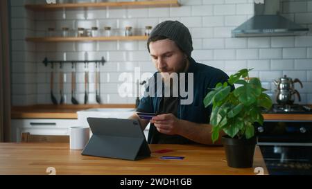 Ein junger Mann mit Beanie, der zu Hause in der Küche sitzt, nimmt seine Kreditkarten aus der Tasche und versucht, die Kreditkarte zu finden, die nicht hat Stockfoto