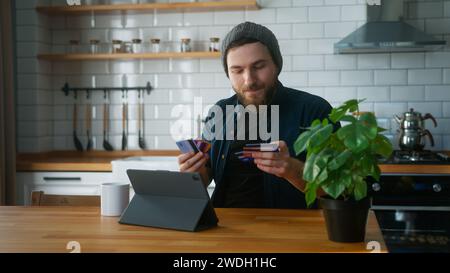 Ein ängstlicher Mann mit Beanie, der zu Hause in der Küche sitzt, nimmt seine Kreditkarten aus der Tasche und versucht, die Kreditkarte zu finden, die er hat Stockfoto