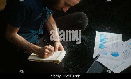 High-Adler-Aufnahme eines jungen Erwachsenen mit langen Haaren, der auf dem Teppich auf dem Boden sitzt und Notizen auf dem Tablet-Computer schreibt Stockfoto