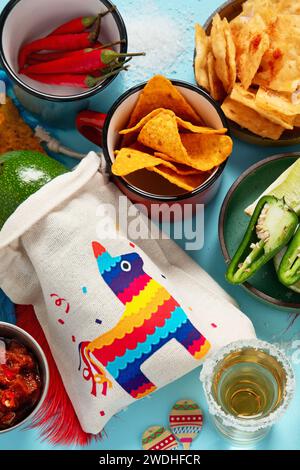 Cinco-de-Mayo-Konzept. Traditionelles Essen Nacho Chips Salsa Sauce Chili Tequila mit Salz Limette Sombrero Serape Kakteen und Maracas auf blauem Hintergrund. Stockfoto