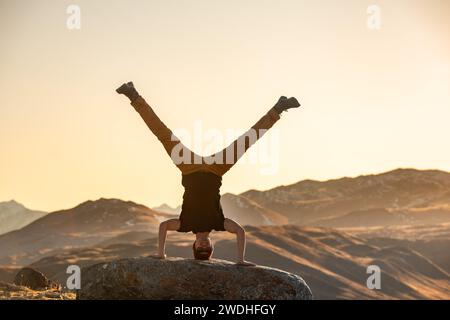 Ein aktiver junger Mann steht kopfüber auf einem großen Felsen in den Bergen des Sonnenuntergangs Stockfoto