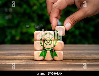 Business Quality and Service Success Konzept. Das letzte Stück des kleinen Holzwürfelblocks von Hand auf Puzzletapel mit großem grünen Garantiesymbol legen Stockfoto