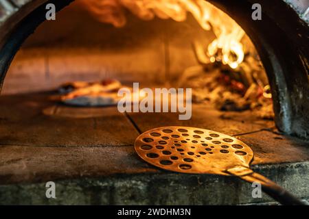 Traditioneller Ofen zum Backen von Pizza mit brennendem Holz und Schaufel. Eine Pizzaschaufel aus Edelstahl, die am Rand eines Backofens platziert wird. Stockfoto