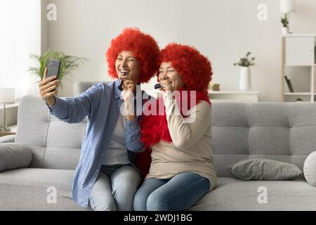 Fröhliche Mutter und erwachsene Tochter in Clown Perücken mit Smartphone Stockfoto