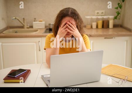 Eine weinende Frau mit Laptop sitzt an einem Tisch in einer Küche. Eine erwachsene Geschäftsfrau arbeitet von zu Hause aus, in einem entfernten Büro Stockfoto