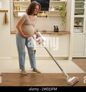 Die werdende Mutter räumt die Küche mit einem kabellosen Vakuum auf. Die schwangere Frau benutzt einen kabellosen Staubsauger, um die Küche zu reinigen. Stockfoto