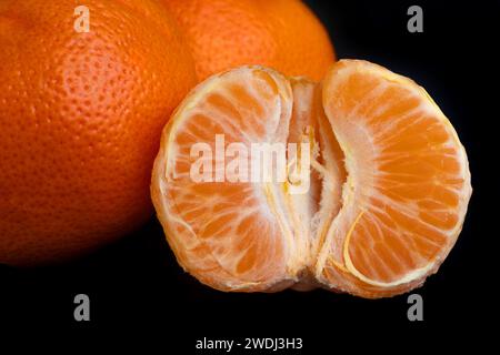 Die Hälfte einer geschälten Mandarine vor dem Hintergrund anderer ganzer Mandarinen. Der Hintergrund des Fotos ist schwarz. Gesunde Ernährung. Saftige Früchte. Zitrusfrüchte im Stockfoto