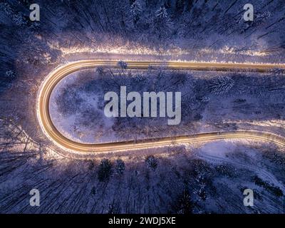 Autoscheinwerfer beleuchten eine kurvenreiche Winterstraße nach Einbruch der Dunkelheit. Lange Belichtungszeit. Transport im Winter bei Nacht. Stockfoto