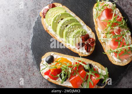 Drei verschiedene offene Sandwiches mit Rucola, Marmelade, sonnengetrockneten Tomaten, Avocado, Frischkäse, Oliven und gebratene Paprika Nahaufnahme auf einem Schieferbrett o Stockfoto