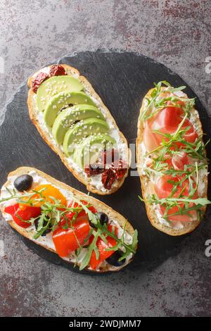 Gemischte Bruschetta-Sandwiches mit Rucola, Schinken, sonnengetrockneten Tomaten, Avocado, Frischkäse, Oliven und gebratene Paprika Nahaufnahme auf einem Schieferbrett auf t Stockfoto