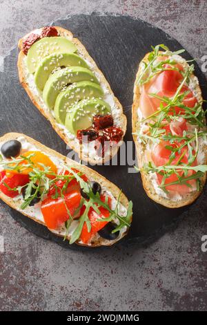 Bruschetta-Sandwiches mit Rucola, Schinken, sonnengetrockneten Tomaten, Avocado, Frischkäse und gebratenen Paprika in Nahaufnahme auf einem Schieferbrett auf dem Tisch. Stockfoto