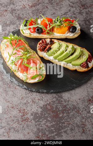 Drei verschiedene offene Sandwiches mit Rucola, Marmelade, sonnengetrockneten Tomaten, Avocado, Frischkäse, Oliven und gebratene Paprika Nahaufnahme auf einem Schieferbrett o Stockfoto