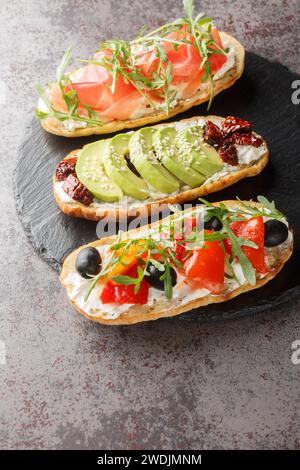 Bruschetta-Sandwiches mit Rucola, Schinken, sonnengetrockneten Tomaten, Avocado, Frischkäse und gebratenen Paprika in Nahaufnahme auf einem Schieferbrett auf dem Tisch. Stockfoto