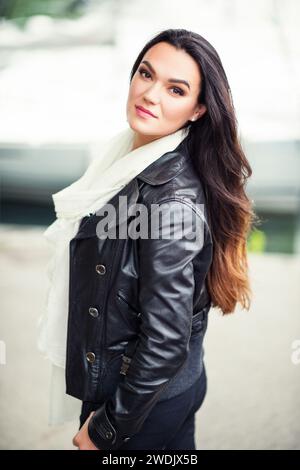 Außenporträt einer schönen brünetten Frau mit langen Haaren, die großen weißen Kaschmirschal und eine schwarze Lederjacke trägt Stockfoto