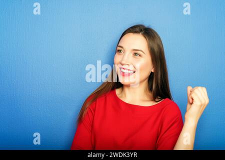 Studio-Aufnahme einer jungen 25-30-jährigen Frau mit rotem Lippenstift und langen dunklen Haaren in rotem Kleid, die an einer blauen Wand steht Stockfoto