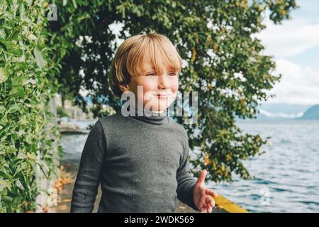 Außenporträt eines lustigen kleinen Jungen, der neben dem wunderschönen Genfer See spaziert, mit grauem Rollkragenhemd Stockfoto