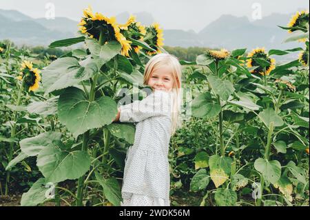 Entzückendes kleines 3-4-jähriges Mädchen, das mit riesigen Sonnenblumen auf einem Feld spielt. Glückliche Kindheit auf dem Land. Herbstmode für Kinder Stockfoto