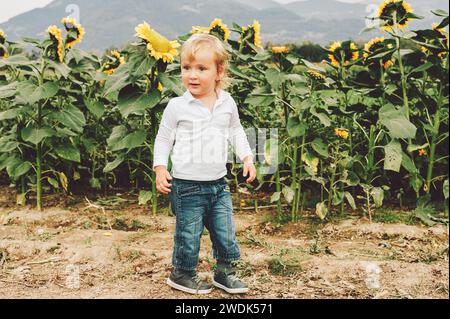 Entzückender kleiner 1-2-jähriger Junge, der mit riesigen Sonnenblumen auf einem Feld spielt. Glückliche Kindheit auf dem Land. Stockfoto