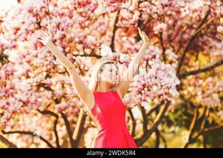 Schöne glückliche junge Frau, die einen warmen sonnigen Tag im Frühlingsgarten genießt, in hellrotem Kleid, die Arme weit offen Stockfoto