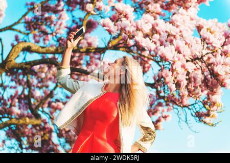 Junge, schöne blonde Frau, die Selfie gegen blühenden Magnolienbaum feiert Stockfoto