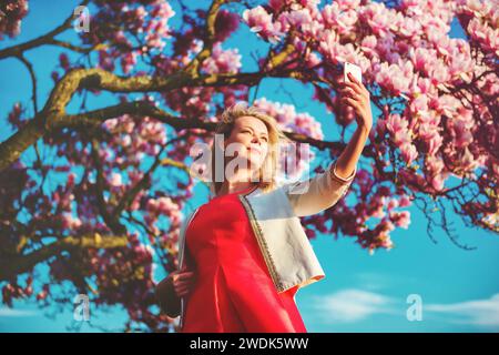 Junge, schöne blonde Frau, die Selfie gegen blühenden Magnolienbaum feiert Stockfoto