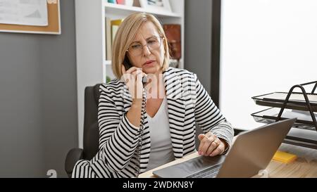 Eine reife Frau in einem Büro, die am Telefon spricht und an einem Laptop arbeitet und eine geschäftige berufliche Umgebung darstellt. Stockfoto