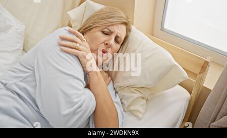 Eine reife kaukasische Frau liegt im Bett mit einem schmerzhaften Ausdruck, der ihre Schulter umklammert, was für Unwohlsein oder Krankheit in Innenräumen steht. Stockfoto