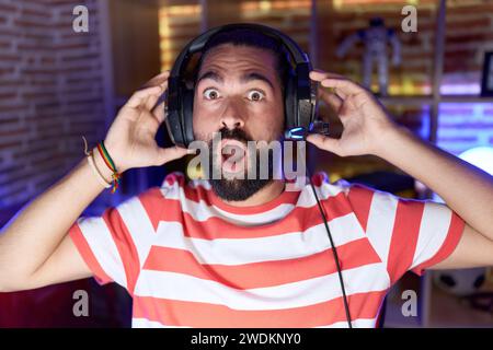 Hispanischer Mann mit Bart, der Videospiele mit Kopfhörern spielt, ängstlich und schockiert vor Überraschung und staunendem Ausdruck, Angst und aufgeregtem Gesicht. Stockfoto