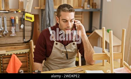 Ein junger hispanischer Mann mit Bart spricht am Telefon in einer Holzwerkstatt, trägt eine Schutzschürze. Stockfoto